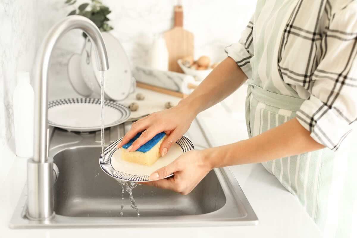 Braccia e mani di donna che insaponano piatto con la spugna nel lavandino